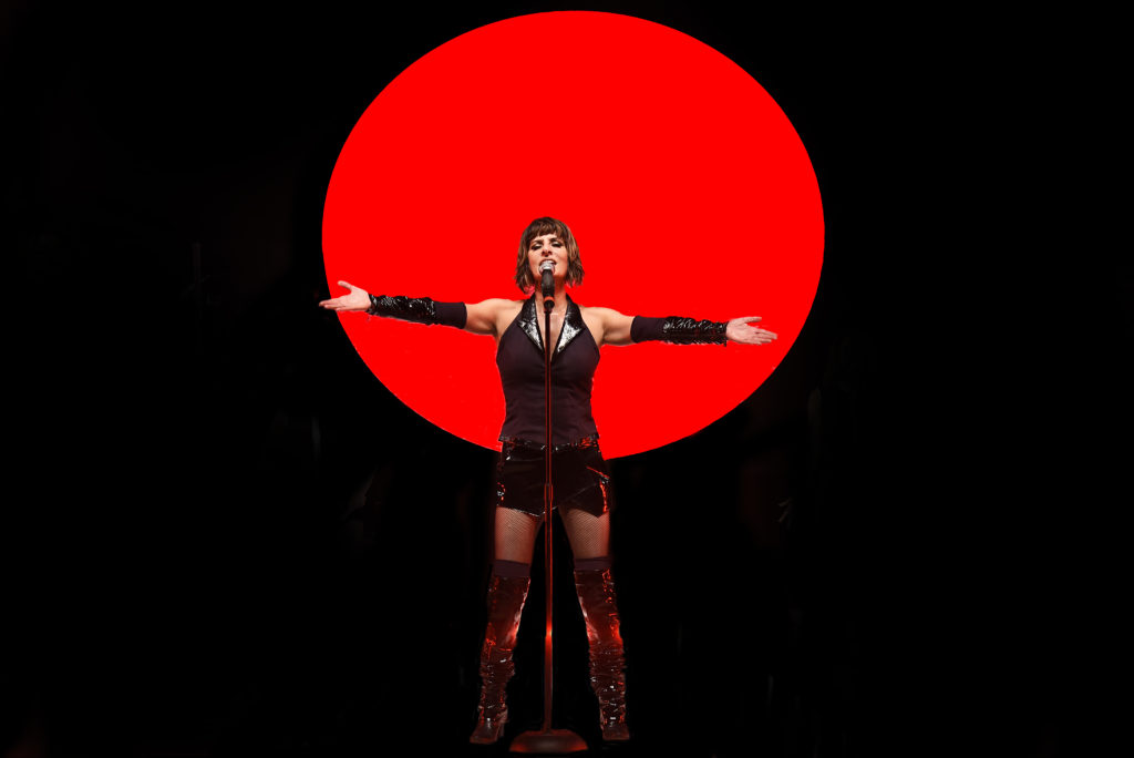 Artista Fernanda Abreu de braços abertos cantando em cima de um palco com fundo preto e um círculo vermelho ao centro. 